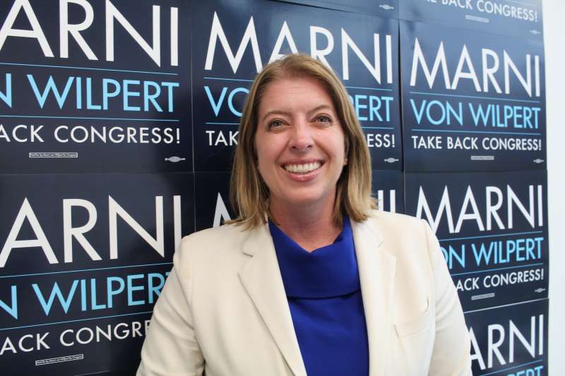 Marni von Wilpert smiles as she stands with shoulder-length blonde hair in front of her campaign signs, wearing a cream blazer and royal blue shirt. Signs behind her say “Marni von Wilpert” and “Take back Congress.”