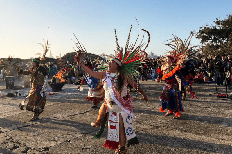 Aztec dancers in feathered headdresses perform at a sunrise ceremony with the San Francisco skyline in the background.